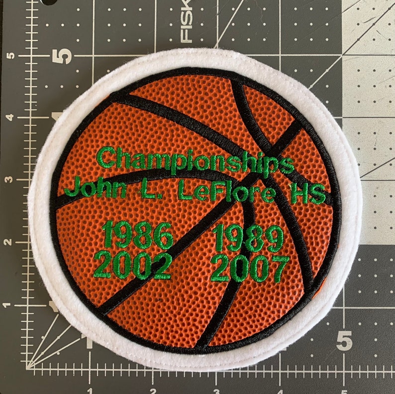 May include: A white and orange basketball patch with black stitching and green text. The text reads "Championships John L. Leflore HS 1986 1989 2002 2007".