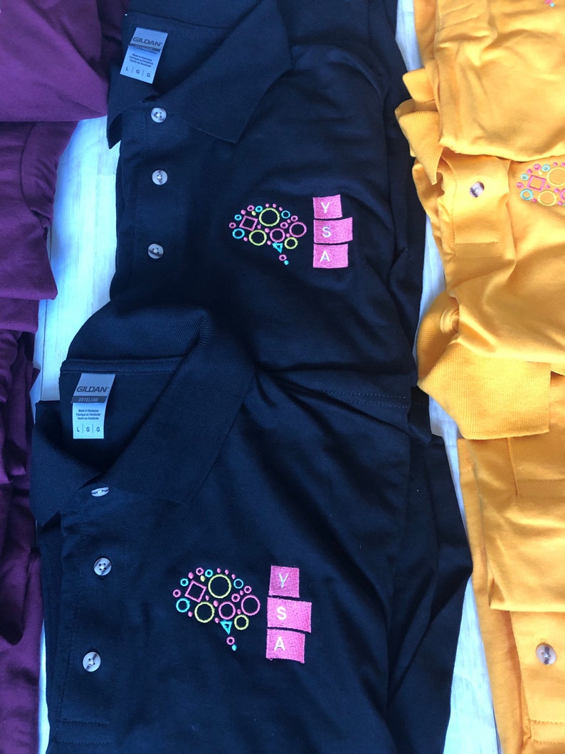 May include: Two black polo shirts with a colorful brain graphic and the letters YSA embroidered on the right chest. The shirts are folded and stacked on a white surface.