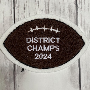 May include: An oval-shaped brown patch with a white border, featuring a white embroidered football lace design at the top and the white text DISTRICT CHAMPS 2024 centered on the textured brown surface of the patch.
