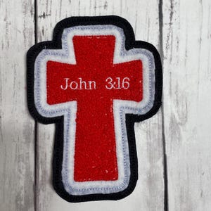 May include: A cross-shaped patch featuring a vibrant red center, a white border, and a thick black outer edge. The text John 3:16 is embroidered in white thread across the center of the red cross, creating a clear and bold religious design.