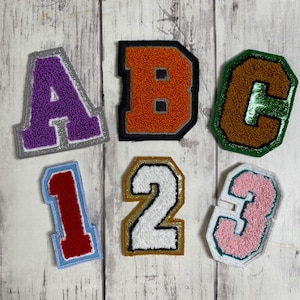 May include: A collection of six varsity-style patches featuring the letters A, B, and C, and the numbers 1, 2, and 3. The letter A is purple with a silver border, B is orange with a black border, and C is brown with a green border. The numbers are red, white, and pink.