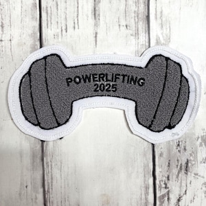 Pode incluir: Um emblema bordado cinza em forma de haltere com contornos pretos e uma borda branca. O centro do haltere apresenta o texto bordado em preto POWERLIFTING 2025. O emblema tem uma superfície texturizada e um design de barra de pesos.