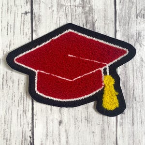 Peut inclure: Un écusson en forme de toque de diplômé avec un dessus carré rouge et une base rouge, bordé de fil noir. Un pompon jaune texturé pend sur le côté droit. Le motif est réalisé dans un style brodé dense et en relief avec une bordure noire.