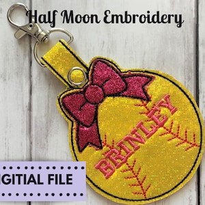 Op de afbeelding: Een gele softbal met een roze strik en de naam "BRITTNEY" erop geborduurd. De softbal is bevestigd aan een zilveren sleutelhanger.