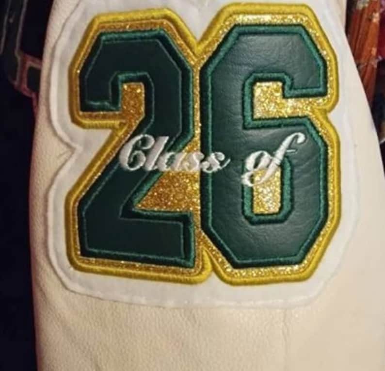 K&ouml;nnte beinhalten: Eine wei&szlig;e Lederjacke mit einem gr&uuml;nen und goldenen Aufn&auml;her, auf dem "Class of 26" steht.