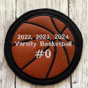 May include: A black and orange embroidered basketball patch with the text "2022, 2023, 2024 Varsity Basketball #0".