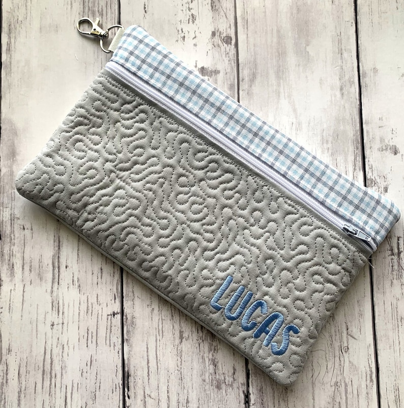 May include: A gray zippered pouch with a blue and white plaid fabric lining. The pouch has a gray quilted pattern and the name "Lucas" is embroidered on the front in blue thread.