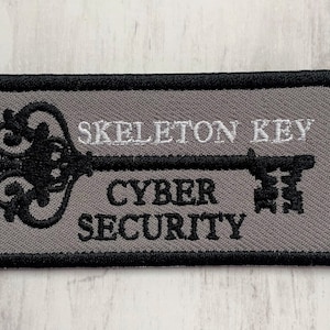 K&ouml;nnte beinhalten: Ein grauer gestickter Aufn&auml;her mit einem schwarzen Skelettschl&uuml;ssel-Design. Der Text "Skeleton Key" und "Cyber Security" ist in Wei&szlig; auf den Aufn&auml;her gestickt.