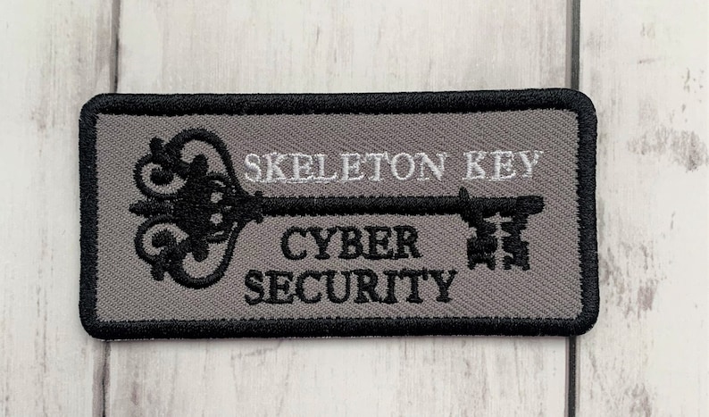 K&ouml;nnte beinhalten: Ein grauer gestickter Aufn&auml;her mit einem schwarzen Skelettschl&uuml;ssel-Design. Der Text "Skeleton Key" und "Cyber Security" ist in Wei&szlig; auf den Aufn&auml;her gestickt.