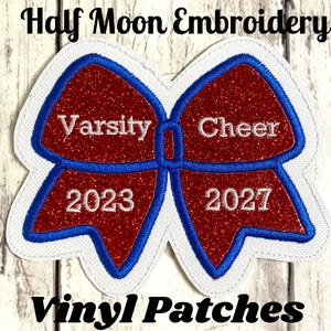May include: A red glittery bow-shaped patch with blue embroidered borders and white stitching. The text Varsity Cheer is embroidered in white, with the years 2023 and 2027 featured below. The patch is set against a white fabric backing.