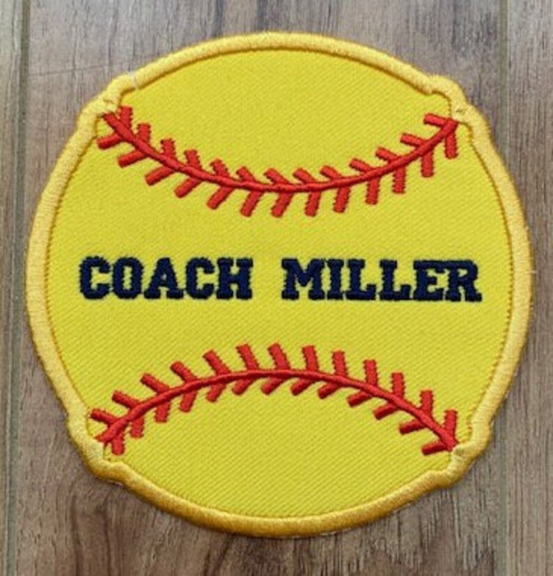May include: Yellow softball-shaped patch with red stitching and the text "COACH MILLER" in navy blue.