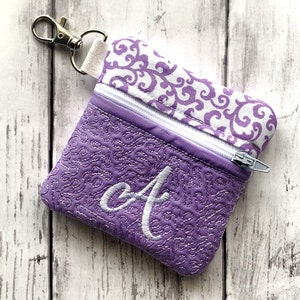 May include: A small purple zippered pouch with a white swirl pattern on the top flap and a white embroidered letter "A" on the front. The pouch has a silver zipper and a silver key ring.