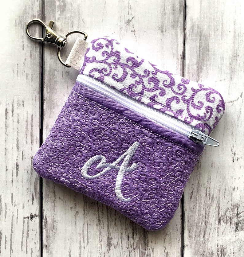 May include: A small purple zippered pouch with a white swirl pattern on the top flap and a white embroidered letter "A" on the front. The pouch has a silver zipper and a silver key ring.