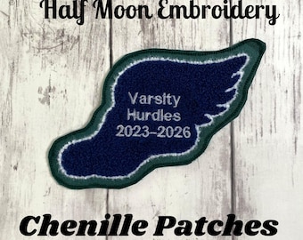 May include: A chenille patch in the shape of a wing, with the text "Varsity Hurdles 2023-2026" embroidered in white on a navy blue background. The patch is outlined in green and white. The words "Half Moon Embroidery" and "Chenille Patches" are also visible.