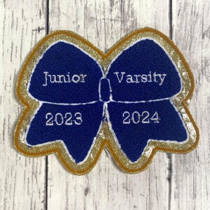 May include: A blue bow-shaped patch with a gold glitter border. The left side features the white embroidered text Junior and 2023, while the right side features the white embroidered text Varsity and 2024. The patch has a textured surface.