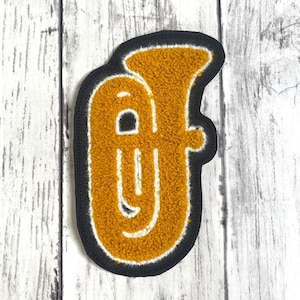 May include: A mustard yellow and white embroidered patch in the shape of a tuba. The musical instrument design features a textured, fuzzy surface with a thick black border outlining the entire shape, creating a distinct and bold visual contrast.