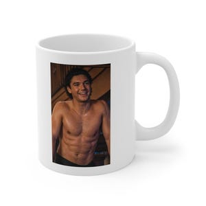 Puede incluir: Taza de café de cerámica blanca con una imagen impresa de una persona sonriente. La persona está sin camisa y de pie frente a un fondo de madera. La taza tiene un asa estándar y un acabado brillante.