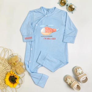 May include: Light blue baby sleeper with a helicopter graphic and the text "I'm Heli-cute." The name "Hayden" is printed on the sleeve. The sleeper is surrounded by baby accessories, including sunglasses and slippers.