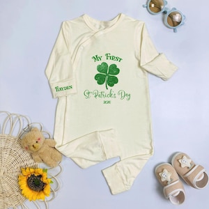 May include: Cream-colored baby sleeper with long sleeves and attached feet. The front features a green four-leaf clover and the text "My First St. Patrick's Day 2026." The sleeve has the name "Hayden" printed on it. Includes matching baby shoes and sunglasses.