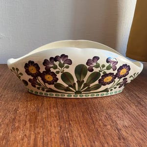 May include: An oval-shaped cream-coloured ceramic bowl featuring a hand-painted floral pattern of purple flowers with yellow centres and green leaves. The rim has a scalloped edge, and the base is decorated with a row of small green dots.