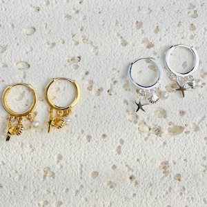 May include: Two pairs of hoop earrings with charms. One pair is gold with a starfish, seashell, and pearl charms. The other pair is silver with the same charms.