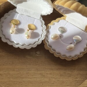 May include: Two pairs of pearl earrings presented in seashell-shaped boxes. One pair has gold-coloured seashell tops, the other silver. Each earring features a teardrop-shaped pearl pendant. The boxes are displayed on a wooden tray.