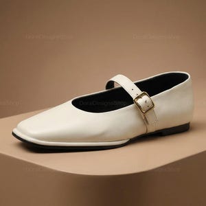 May include: Off-white leather Mary Jane flat shoe with a square toe and a strap across the top of the foot. The strap fastens with a gold-colored buckle. The shoe has a black sole.