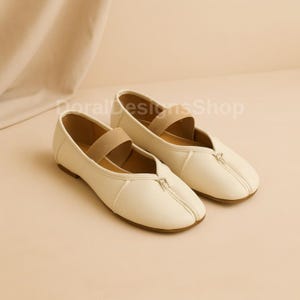May include: A pair of off-white flat shoes featuring a split-toe design, a central vertical seam, and a beige elastic strap across the instep. The shoes have a smooth finish and a low profile. The text DoralDesignsShop is visible in the background.