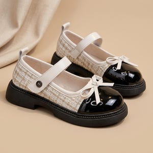 Könnte beinhalten: Ein Paar Mary-Jane-Schuhe mit einem Obermaterial aus beigefarbenem und weißem Tweed-Stoff, einer abgerundeten Zehenkappe aus glänzendem schwarzem Lackleder, einer Zierschleife und einem Riemen mit Metallknopfverschluss auf einer dicken schwarzen Gummisohle.