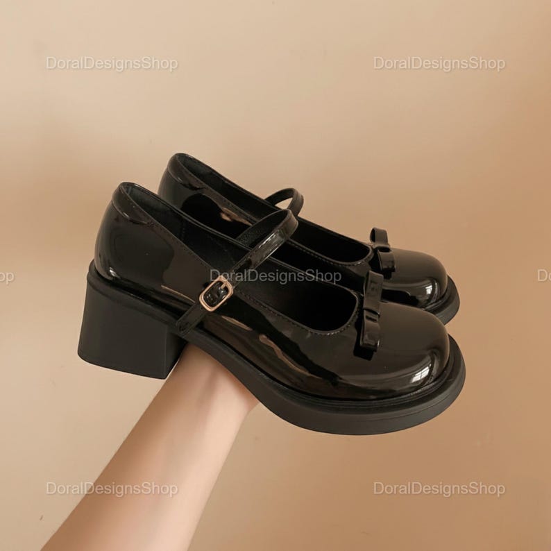 May include: Pair of black patent leather Mary Jane shoes with a chunky heel and a strap with a buckle closure. Each shoe has a small bow on the toe. The shoes are on a neutral background.