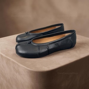 May include: A pair of black leather ballet flats with a rounded toe and a small bow detail. The shoes are displayed on a neutral-toned surface, showcasing their sleek design and minimalist aesthetic. The flats appear to be unworn.