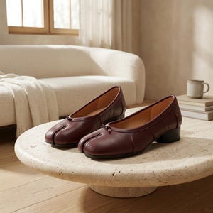 May include: A pair of dark burgundy leather split-toe ballet flats featuring a low block heel, a rounded topline, and a small decorative bow detail at the center of the vamp. The shoes have a smooth finish and a distinct cleft design at the toe.