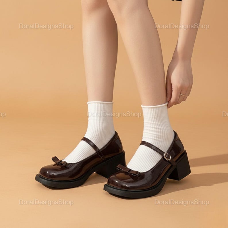 May include: Dark brown Mary Jane style shoes with a bow detail and a block heel. The shoes have a glossy finish and a strap with a buckle. The shoes are worn with white socks.