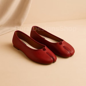 May include: A pair of red ballet flats with a split-toe design. The shoes have a low heel and a rounded toe. The interior is white, and the shoes are made of a smooth material. The shoes are displayed on a neutral background.