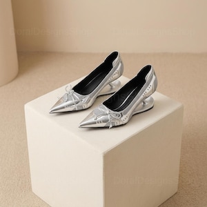 May include: A pair of silver pointed-toe pumps with a unique heel design. The shoes feature a mesh overlay on the toe and a small bow detail. The pumps are displayed on a white cube.