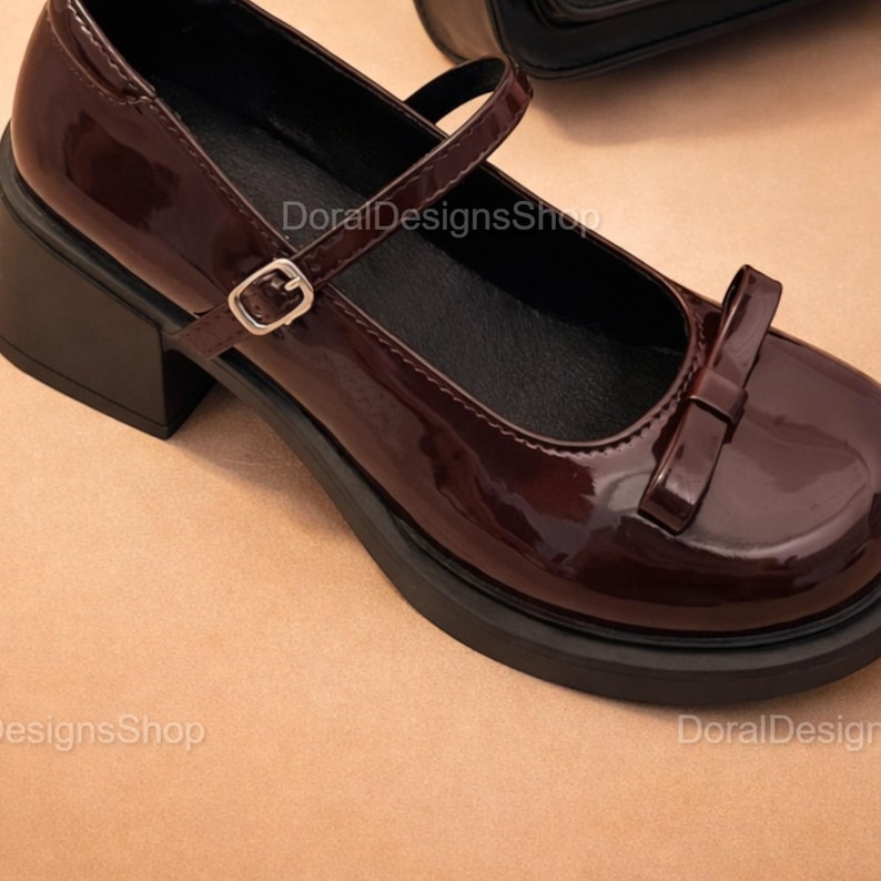 May include: Burgundy Mary Jane style shoe with a bow detail and a strap with a buckle. The shoe has a block heel and a platform sole. The shoe is patent leather and has the text "DoralDesignsShop".