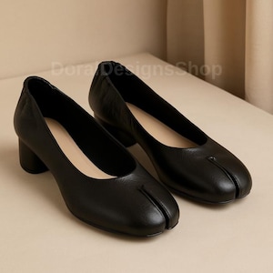 Può includere: Un paio di scarpe in pelle nera con design a punta divisa e tacco basso a blocco. Le scarpe presentano una silhouette liscia e arrotondata con una distinta fessura verticale nella zona della punta anteriore. La fodera interna è beige. DoraDesignsShop.