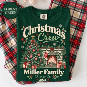 May include: A forest green t-shirt featuring a graphic of a Christmas tree, a fireplace with stockings, and wrapped gifts. The text reads Christmas Crew, Miller Family, Chicago, IL, and 2026. The design includes snowflakes, holly, and ornaments.