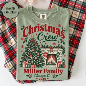 May include: A sage green t-shirt featuring a graphic with the text Christmas Crew, Miller Family, Chicago, IL, 2026. The design includes a decorated Christmas tree, a fireplace with stockings, and wrapped gifts in red, white, and green colors.