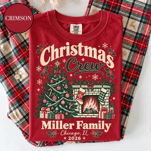 May include: A crimson t-shirt featuring a graphic of a decorated Christmas tree, a fireplace with stockings, and the text "Christmas Crew", "Miller Family", "Chicago, IL", and "2026". The shirt has a Comfort Colors label at the inner neckline.