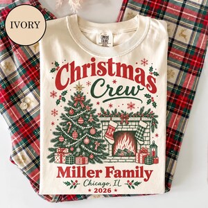 May include: An ivory t-shirt featuring a graphic of a Christmas tree, a fireplace with stockings, and gifts. The text reads: Christmas Crew, Miller Family, Chicago, IL, 2026. The design includes red and green accents, snowflakes, and holly leaves.