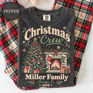 May include: A charcoal gray t-shirt featuring a graphic print with the text "Christmas Crew", "Miller Family", "Chicago, IL", and "2026". The design includes a decorated Christmas tree, a fireplace with stockings, and wrapped gift boxes.