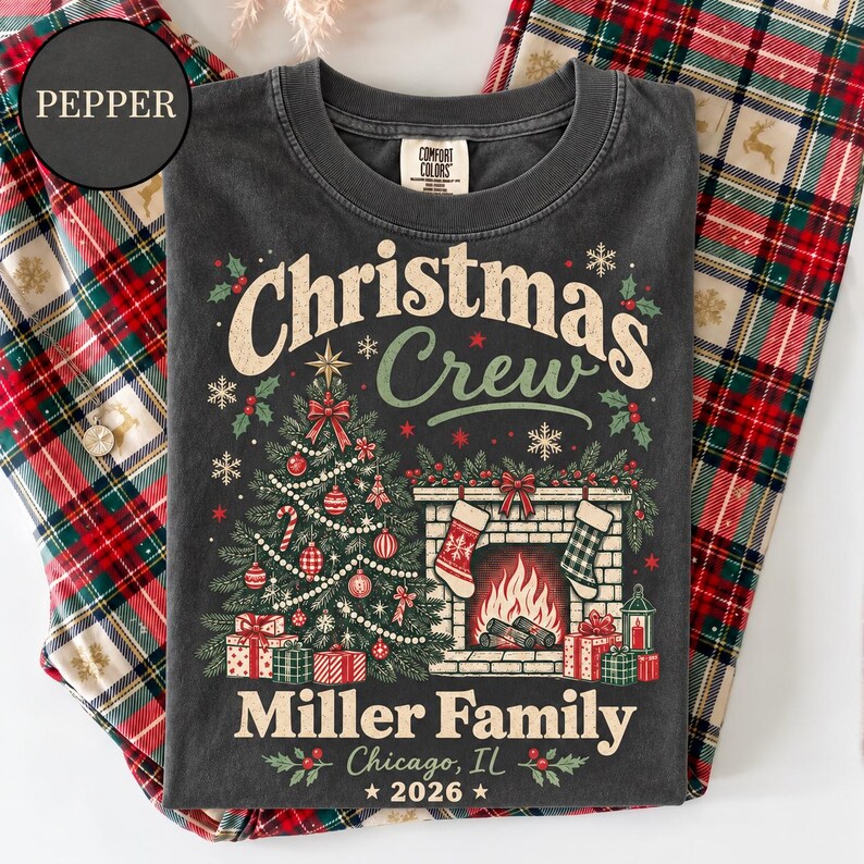 May include: A charcoal gray t-shirt featuring a graphic print with the text "Christmas Crew", "Miller Family", "Chicago, IL", and "2026". The design includes a decorated Christmas tree, a fireplace with stockings, and wrapped gift boxes.