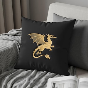 May include: Black square throw pillow featuring a gold embroidered dragon design. The dragon is depicted with outstretched wings and a fiery breath. The pillow is placed on a gray sofa, adding a touch of fantasy decor.