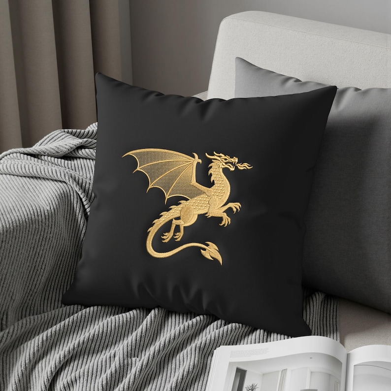 May include: Black square throw pillow featuring a gold embroidered dragon design. The dragon is depicted with outstretched wings and a fiery breath. The pillow is placed on a gray sofa, adding a touch of fantasy decor.