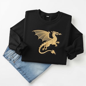 May include: Black sweatshirt with a gold embroidered dragon design. The dragon is depicted with outstretched wings and a fiery breath. The sweatshirt is paired with blue jeans, creating a casual outfit.