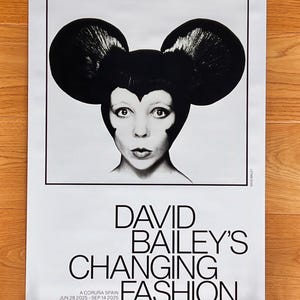 Peut inclure: Une affiche d'exposition rectangulaire présentant un portrait en noir et blanc d'une personne aux cheveux volumineux et arrondis en forme d'oreilles. Le texte indique : DAVID BAILEY'S CHANGING FASHION, A CORUÑA SPAIN, JUN 28 2025 - SEP 14 2025, WWW.THEMOPFOUNDATION.ORG.