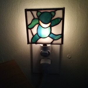May include: A stained glass nightlight featuring a green bear with a white outline. The nightlight is plugged into a standard electrical outlet.