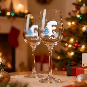 Könnte beinhalten: Zwei klare Weingläser mit personalisierten Wintermotiven. Ein Glas zeigt den Namen "Faith" mit dem Buchstaben "F" und einem Weihnachtsbaum-Design. Das andere Glas hat den Namen "Cindy". Die Gläser stehen auf einem Tisch mit Weihnachtsdekorationen.
