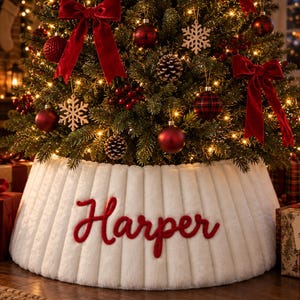 Op de afbeelding: Een witte kerstboomrok van imitatiebont met een verticale ribbelstructuur en de naam Harper geborduurd in rode schrijfletters. De rok ligt aan de voet van een kerstboom die versierd is met rode strikken en kerstballen.